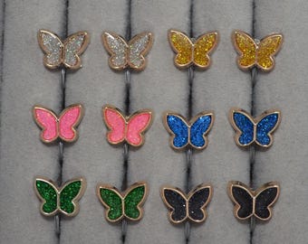 Peut inclure: Une collection de douze bagues en forme de papillon disposées sur trois rangées. Chaque bague présente un papillon pailleté de différentes couleurs, notamment argent, jaune, rose, bleu, vert et noir, monté sur un anneau métallique fin.