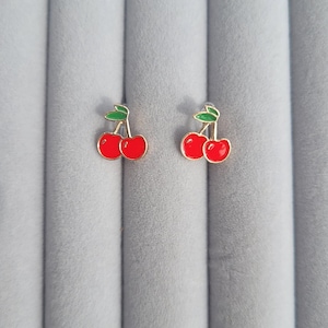 Peut inclure: Une paire de boucles d'oreilles cerises. Chaque boucle d'oreille présente un motif de cerise rouge avec des feuilles vertes et des accents dorés. Les boucles d'oreilles sont posées sur un fond gris.