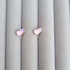 Peut inclure: Deux paires de boucles d'oreilles à clip en forme de cœur. Chaque boucle d'oreille présente un cœur rose associé à un cœur violet, monté sur du métal doré. Les boucles d'oreilles sont présentées sur un fond gris texturé.