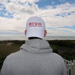 Può includere: Un berretto da baseball bianco con la scritta "NUTS ACK" in rosso, con un disegno a forma di cuore. Il testo "THE NANTUCKET NUT COMPANY" è sotto. La persona indossa una felpa con cappuccio grigia, con vista sul mare e sul cielo sullo sfondo.