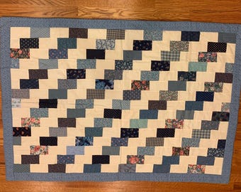 May include: A handmade quilt with a blue and cream color scheme. The quilt features a repeating pattern of staggered blocks in various shades of blue and floral prints, bordered by a light blue fabric.