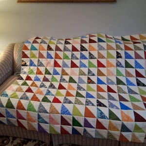 May include: A handmade quilt with a patchwork design of colorful triangles in shades of red, blue, green, orange, and purple. The quilt is draped over a sofa, showcasing its geometric pattern and vibrant colors. The quilt is a cozy home decor item.