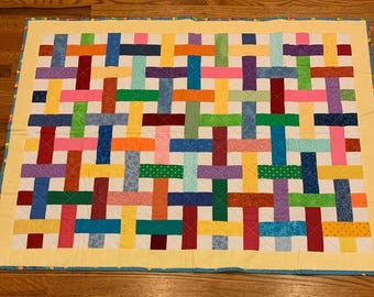 May include: A colorful handmade quilt with a woven pattern. The quilt features a variety of bright colors including red, blue, green, yellow, and purple. The quilt is bordered with a yellow fabric and a blue dotted trim.