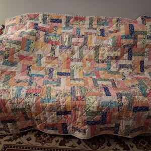 May include: A colorful patchwork quilt with a geometric design, featuring rectangles in various pastel shades of pink, blue, yellow, and green. The quilt is draped over a sofa, showcasing its intricate stitching and floral patterns.