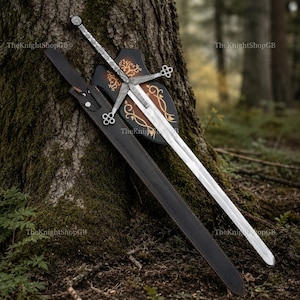 May include: A silver-toned metal sword with a cross-shaped hilt featuring intricate knotwork, accompanied by a black leather scabbard and a dark brown wooden plaque with gold-coloured filigree designs. The text TheKnightShopGB is visible.