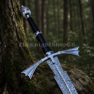 May include: A sword with a patterned Damascus steel blade, a black grip, and a polished silver-coloured pommel and crossguard. The crossguard features flared, textured ends. The text TheKnightShopGB is visible across the centre of the blade.