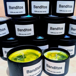 May include: A collection of black tin candles with white labels that read "Banditos 100% soy wax candle." The candles have a yellow and green wax interior. The candles are stacked on top of each other.