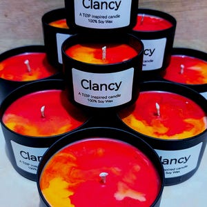 Op de afbeelding: Een verzameling zwarte kaarsen in containers met rood en geel was. Elke kaars heeft een wit etiket met de tekst "Clancy A TOP inspired candle 100% Soy Wax". De kaarsen zijn in een piramidevorm gerangschikt.
