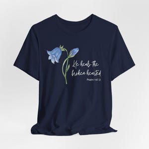 Op de afbeelding: Marineblauw T-shirt met een aquarel illustratie van een blauwe klokjesbloem en de tekst "He heals the broken hearted" met "Psalm 147.3" eronder. Het ontwerp staat op de voorkant.