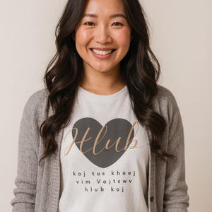 Pode incluir: Uma mulher vestindo um cardigan cinza sobre uma camiseta branca. A camiseta apresenta um coração preto com a palavra "Hlub" em escrita dourada e texto adicional abaixo. A mulher está sorrindo.