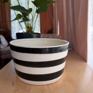 May include: A round ceramic planter featuring a wide, off-white base color with three distinct, horizontal black stripes encircling the exterior. The interior of the bowl-shaped vessel is a smooth, solid off-white finish with a wide, open top rim.