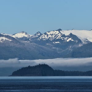 May include: A scenic landscape featuring snow-capped mountains under a clear blue sky. A layer of fog hovers over a dark green island and the calm, dark blue water below.