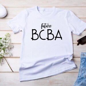 Peut inclure: T-shirt blanc avec l'inscription noire "future BCBA". Le t-shirt est présenté avec des baskets blanches, des lunettes de soleil, une branche de fleurs blanches et un short en jean sur une surface en bois clair.