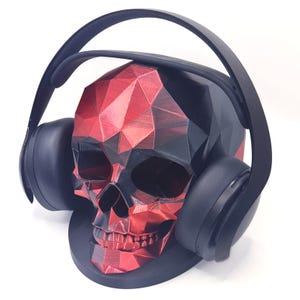 May include: A black and red geometric skull-shaped headphone stand. The skull features a faceted, low-poly design with a metallic red and matte black finish. A pair of solid black over-ear headphones rests securely on the skull's cranium.
