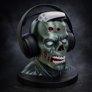 May include: A zombie head-shaped headphone stand in shades of green, red, and white. The stand holds black headphones and a white controller. The base is black and round. The zombie has exposed teeth and red eyes.