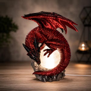 May include: A decorative lamp featuring a glossy, metallic red dragon with black accents, coiled around a glowing, spherical white light. The dragon is perched upon a textured, dark gray rocky base, creating a striking contrast between the light.