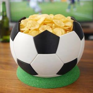 Puede incluir: Un cuenco en forma de balón de fútbol, blanco y negro, lleno de patatas fritas doradas, sobre una base circular verde. El cuenco es de plástico y está diseñado para parecerse a un balón de fútbol. El fondo muestra un televisor y una botella.