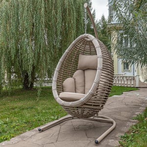 May include: A beige wicker egg-shaped hanging chair with matching cushions. The chair is suspended from a curved metal frame and sits on a stone patio. The background features a green lawn and a large weeping willow tree.