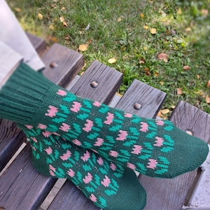 Op de afbeelding: Een paar groene gebreide sokken met een bloemenpatroon in roze en groen. De sokken liggen op een houten bank. De sokken hebben een geribbelde manchet en een patroon op de voet en het been.