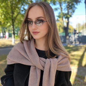 Pode incluir: Óculos de sol sem armação com lentes degradê escuras e hastes metálicas finas, combinados com um suéter de malha bege macio drapeado sobre os ombros e uma blusa preta de manga comprida por baixo, criando um visual em camadas de tons neutros.
