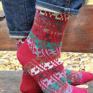 Op de afbeelding: Rode gebreide sokken met een kleurrijk geometrisch patroon. Het patroon bevat groen, wit, bruin en blauw. De sokken worden gedragen met een blauwe spijkerbroek.
