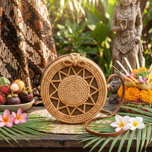 K&ouml;nnte beinhalten: Runde Handtasche aus geflochtenem Rattan mit braunem Lederriemen. Die Tasche hat ein Sonnenstrahl-Design und eine Schleife oben. Sie liegt auf einer Holzoberfl&auml;che mit tropischen Blumen und Bl&auml;ttern.