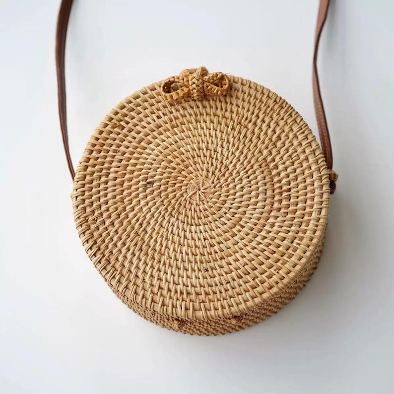 K&ouml;nnte beinhalten: Eine runde, gewebte Korbtasche aus Rattan mit einem braunen Lederriemen. Die Tasche ist hellbraun mit einem spiralf&ouml;rmigen Webmuster und einer kleinen Schleife oben. Der Riemen ist d&uuml;nn.