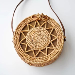 K&ouml;nnte beinhalten: Runde Tasche aus geflochtenem Rattan mit einem braunen Lederriemen. Die Tasche hat ein sternf&ouml;rmiges Muster in der Mitte und eine kleine Schleife oben. Die Tasche hat einen Durchmesser von etwa 25 cm.