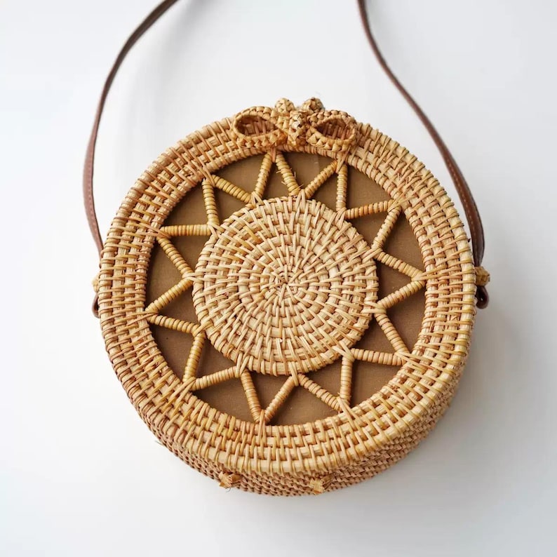 K&ouml;nnte beinhalten: Runde Tasche aus geflochtenem Rattan mit einem braunen Lederriemen. Die Tasche hat ein sternf&ouml;rmiges Muster in der Mitte und eine kleine Schleife oben. Die Tasche hat einen Durchmesser von etwa 25 cm.