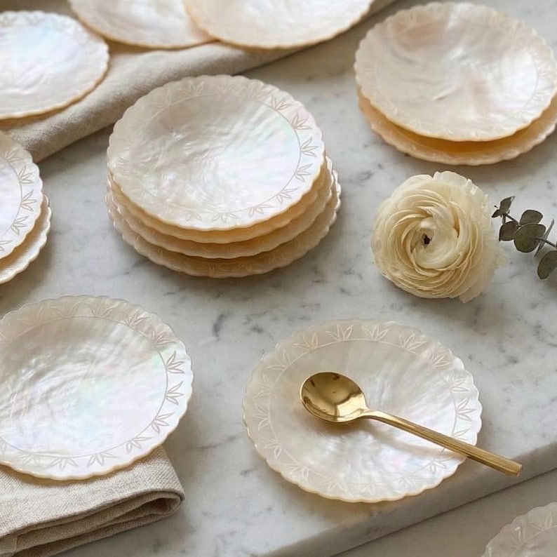 K&ouml;nnte beinhalten: Eine Sammlung schimmernder, runder Untersetzer aus Perlmutt mit einem zarten Blattmuster. Ein goldener L&ouml;ffel liegt auf einem Untersetzer. Eine cremefarbene Blume und ein Zweig mit Gr&uuml;n sind ebenfalls vorhanden.