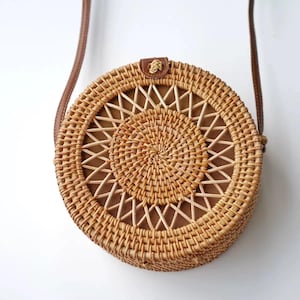 K&ouml;nnte beinhalten: Runde Tasche aus geflochtenem Rattan mit braunem Lederriemen. Die Tasche hat ein kreisf&ouml;rmiges Design mit einem gewebten Muster und einem braunen Innenraum. Der Riemen ist mit einem kleinen Lederst&uuml;ck an der Tasche befestigt. Die Tasche ist naturfarben.