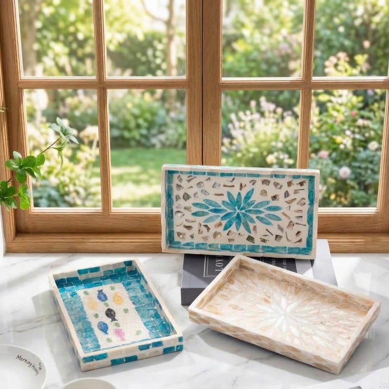 Pode incluir: Tr&ecirc;s bandejas retangulares decorativas com desenhos de mosaico. Uma bandeja apresenta um padr&atilde;o floral azul, outra tem um motivo de peixe e a terceira tem um design floral branco. As bandejas s&atilde;o exibidas em uma superf&iacute;cie branca perto de uma janela.
