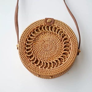 K&ouml;nnte beinhalten: Runde Umh&auml;ngetasche aus geflochtenem Rattan mit braunem Lederriemen. Die Tasche hat ein dekoratives kreisf&ouml;rmiges Muster in der Mitte und einen kleinen Lederaufn&auml;her in der N&auml;he der Riemenbefestigung. Die Tasche hat eine nat&uuml;rliche beige Farbe.