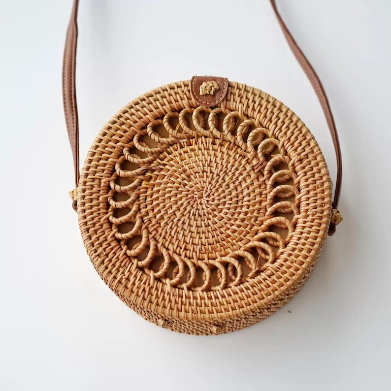 K&ouml;nnte beinhalten: Runde Umh&auml;ngetasche aus geflochtenem Rattan mit braunem Lederriemen. Die Tasche hat ein dekoratives kreisf&ouml;rmiges Muster in der Mitte und einen kleinen Lederaufn&auml;her in der N&auml;he der Riemenbefestigung. Die Tasche hat eine nat&uuml;rliche beige Farbe.