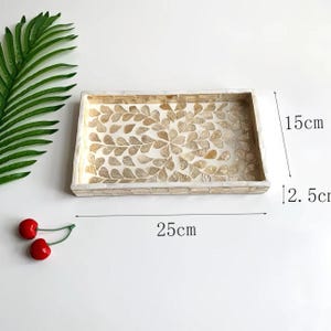 Pode incluir: Bandeja decorativa retangular, com 25 cm de comprimento, 15 cm de largura e 2,5 cm de profundidade. A bandeja apresenta um design de mosaico com um padr&atilde;o de folhas em tons de bege e creme. A borda &eacute; feita de pequenas pe&ccedil;as retangulares.