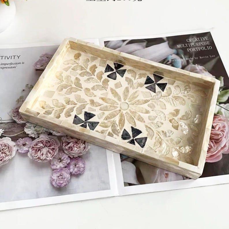 Pode incluir: Bandeja decorativa retangular com um design floral em mosaico em creme, bege e preto. A bandeja &eacute; feita de pequenas pe&ccedil;as irregulares e tem uma borda elevada. A bandeja est&aacute; em cima de uma revista com o texto "CREATIVE MULTIPURPOSE PORTFOLIO".