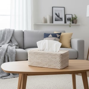 May include: A rectangular tissue box with a woven, beige exterior sits on a wooden coffee table. White tissues are visible at the top. The table is in front of a gray sofa with decorative pillows, creating a cozy living room scene.