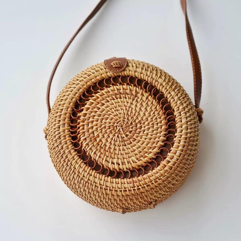 K&ouml;nnte beinhalten: Runde Handtasche aus geflochtenem Rattan mit braunem Lederriemen. Die Tasche hat ein Spiralmuster in der Mitte und einen braunen Lederbesatz. Der Riemen ist mit einem kleinen Lederst&uuml;ck an der Tasche befestigt.