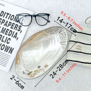 K&ouml;nnte beinhalten: Eine dekorative, irisierende muschelf&ouml;rmige Schale mit silberfarbenem Rand und floralen Akzenten. Die Schale ist 24-28 cm lang und 2-6 cm breit. Eine schwarze Brille und ein gestreifter Hut sind ebenfalls im Bild.