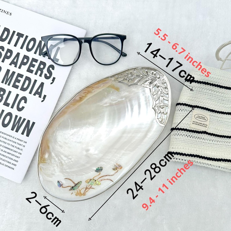 K&ouml;nnte beinhalten: Eine dekorative, irisierende muschelf&ouml;rmige Schale mit silberfarbenem Rand und floralen Akzenten. Die Schale ist 24-28 cm lang und 2-6 cm breit. Eine schwarze Brille und ein gestreifter Hut sind ebenfalls im Bild.