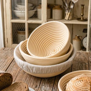 May include: Three natural-colored, round banneton proofing baskets with spiral patterns, nested inside each other. The baskets are made of woven material and are sitting on a wooden surface. A linen liner is visible in the bottom basket.