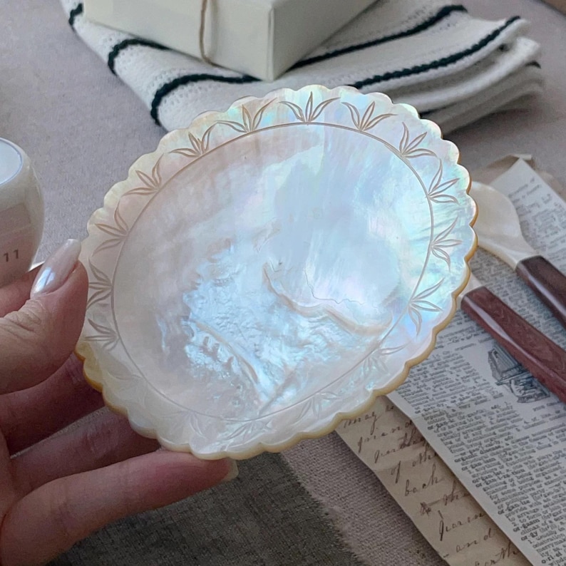 K&ouml;nnte beinhalten: Eine dekorative Platte mit Perlmutt-Finish und gewelltem Rand. Die Platte hat einen schillernden Glanz mit ge&auml;tzten blattartigen Designs am Rand. Die Platte wird von einer Person gehalten. Der Hintergrund umfasst ein wei&szlig;-gr&uuml;n gestreiftes Tuch und Vintage-Papier.