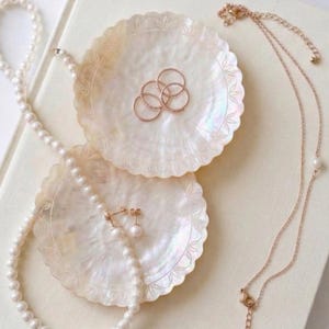 K&ouml;nnte beinhalten: Zwei schimmernde Muschelschalen mit gewelltem Rand halten ros&eacute;goldene Ringe und Perlenohrringe. Eine Perlenkette und eine zarte Halskette aus Ros&eacute;gold mit einem Perlenanh&auml;nger sind ebenfalls vorhanden. Der Schmuck ist auf einer cremefarbenen Oberfl&auml;che angeordnet.