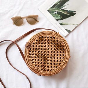 K&ouml;nnte beinhalten: Runde, gewebte Rattan-Handtasche mit braunem Lederriemen und Akzenten. Die Tasche ist hellbraun und hat ein geometrisches Muster. Eine hellbraune Sonnenbrille und eine Zeitschrift sind ebenfalls im Bild.