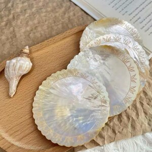 K&ouml;nnte beinhalten: Ein Satz schillernder, runder Untersetzer mit gewelltem Rand und goldenem Blattdesign. Die Untersetzer bestehen aus einem perlmuttartigen Material und werden auf einem Holztablett mit einer Muschel pr&auml;sentiert.