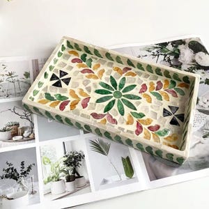 Pode incluir: Bandeja decorativa retangular com um design de mosaico. A bandeja apresenta um padr&atilde;o floral verde central rodeado por formas de folhas coloridas em vermelho, amarelo e verde. A borda &eacute; contornada com pequenas pe&ccedil;as de mosaico verdes.
