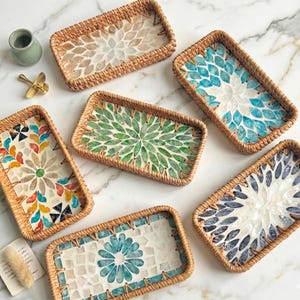May include: A collection of rectangular woven trays featuring mosaic floral patterns. The trays have a natural brown woven border and are displayed on a marble surface. The mosaic designs incorporate shades of blue, green, and neutral tones.