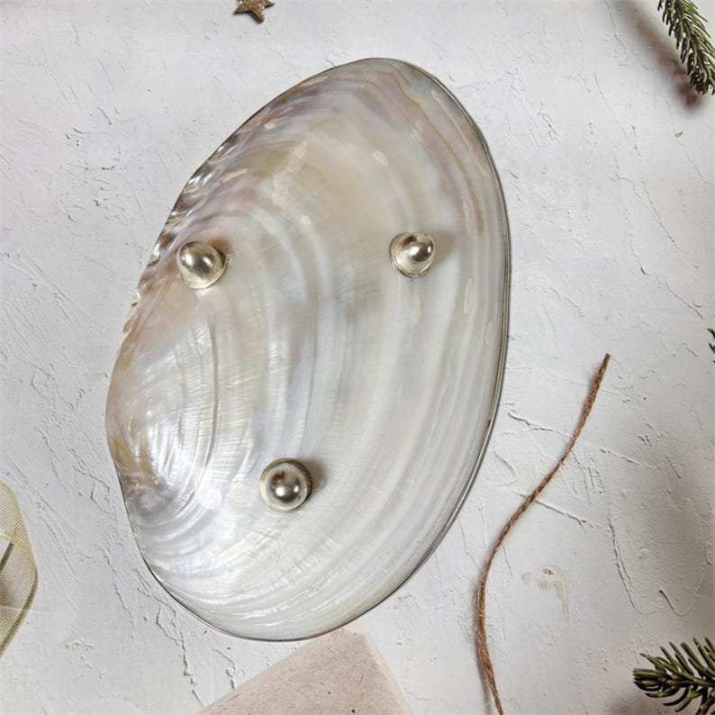 K&ouml;nnte beinhalten: Eine ovale, schillernde Muschelschale mit drei silberfarbenen, kugelf&ouml;rmigen F&uuml;&szlig;en. Die Schale hat einen perlmuttartigen Glanz mit wirbelnden Mustern in Wei&szlig;, Creme und Grau. Ein dekorativer Artikel zur Pr&auml;sentation kleiner Objekte.