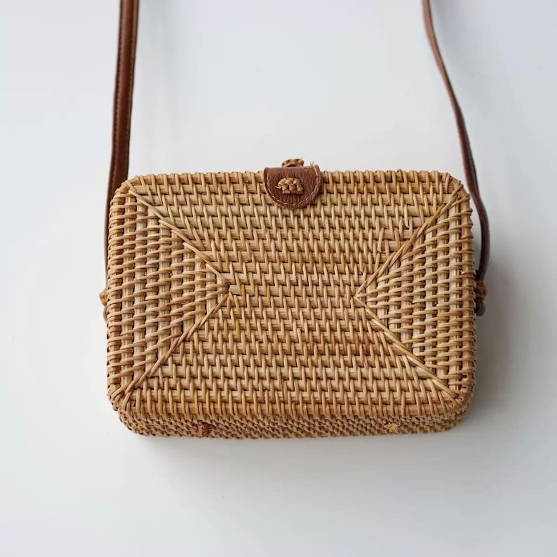 K&ouml;nnte beinhalten: Rechteckige Umh&auml;ngetasche aus geflochtenem Rattan mit braunem Lederriemen und Lederklappenverschluss. Die Tasche ist hellbraun und hat ein gewebtes Muster. Der Riemen ist d&uuml;nn und braun. Die Tasche h&auml;ngt vor wei&szlig;em Hintergrund.