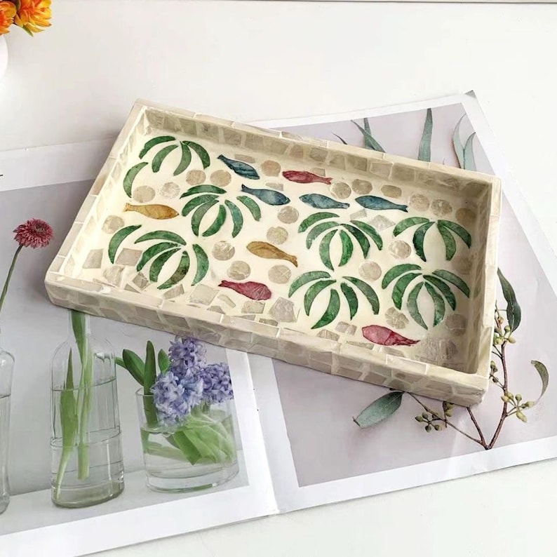 Pode incluir: Bandeja decorativa retangular com um design de mosaico. A bandeja apresenta um fundo creme com palmeiras verdes e peixes coloridos. A borda &eacute; feita de pequenas telhas quadradas.
